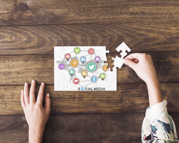 Photographie en vue plongeante (prise du dessus), sur une table en bois brun foncé, avec des veines du bois bien visibles et un aspect naturel, légèrement rustique.  Au centre de l’image se trouve un puzzle rectangulaire presque terminé. Le puzzle représente une carte du monde stylisée, en gris, recouverte d’icônes circulaires colorées reliées entre elles par des lignes pointillées, symbolisant un réseau.  Les icônes correspondent à des réseaux sociaux et outils numériques, reconnaissables par leurs couleurs et logos :  Twitter (oiseau blanc sur fond bleu),  Facebook (F blanc sur fond bleu),  Pinterest (P blanc sur fond rouge),  Google+ (G blanc sur fond orange),  YouTube (icône rouge),  d’autres pictogrammes de profils humains et de partage, en vert, violet, orange et rouge.  Sous la carte, le texte « SOCIAL MEDIA » est visible en lettres capitales noires, partiellement intégré au puzzle.  Deux mains humaines sont visibles :  à gauche, une main posée à plat sur la table, doigts légèrement écartés, immobile ;  à droite, une main tenant une pièce de puzzle blanche, encore non placée, située juste à côté de l’espace vide correspondant dans le puzzle.  La scène évoque l’idée de construction, d’assemblage et de stratégie autour des réseaux sociaux et du numérique, avec une métaphore claire : tout n’est pas encore en place, mais presque. Il manque juste la dernière pièce… comme souvent avec les réseaux sociaux.